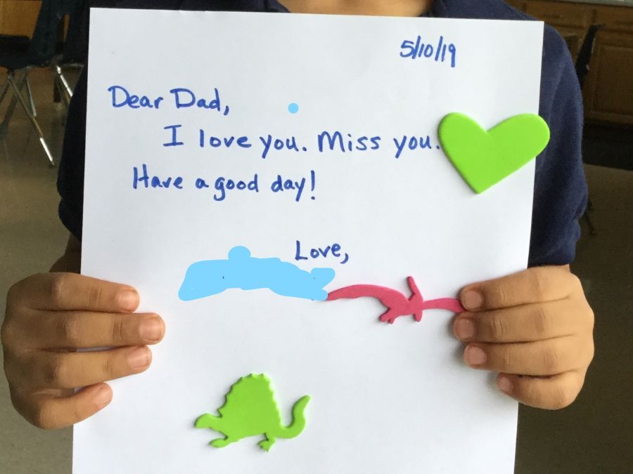 Child holding up letter reading, "Dear Dad, I love you. Miss you. Have a good day!"